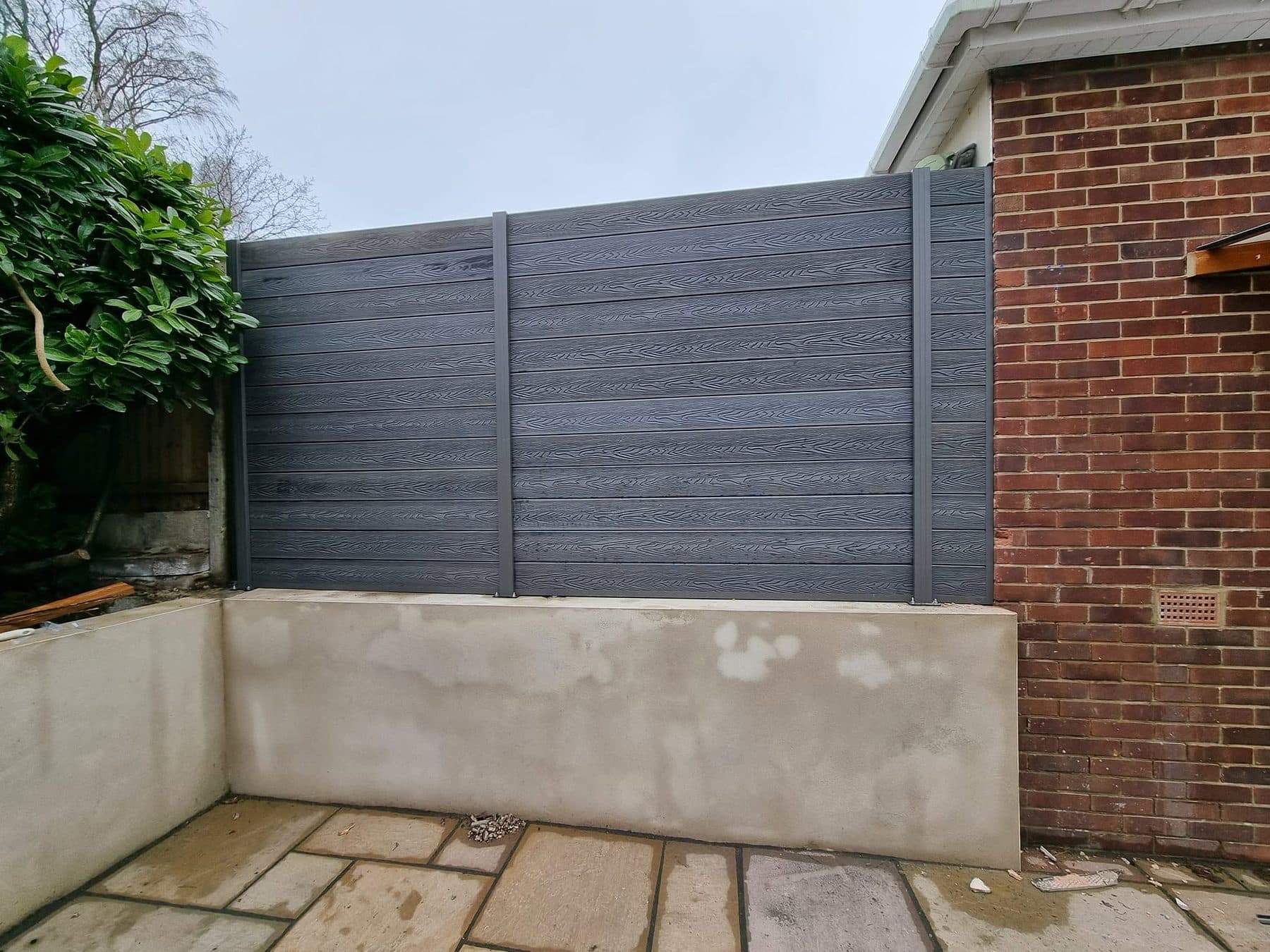 Grey composite fence panels fixed on top of a new rendered retaining wall, sandstone patio in front