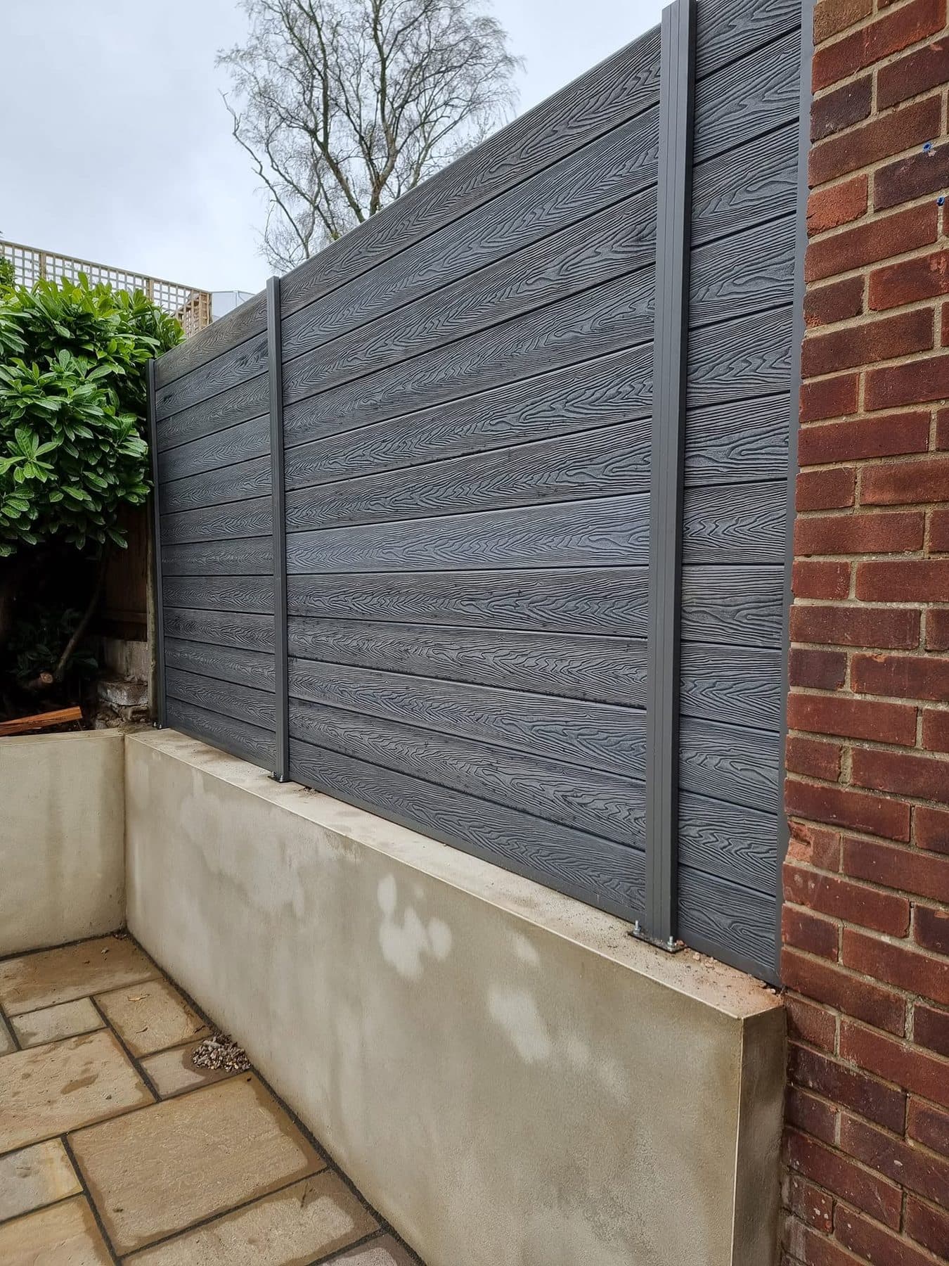 Angled view of grey composite fencing on a rendered block wall, with base-plated posts and a sandstone patio