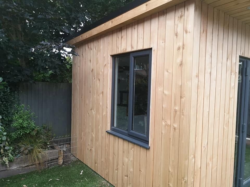 Side of a timber-clad garden room with an anthracite grey window, on artificial grass beside a grey fence