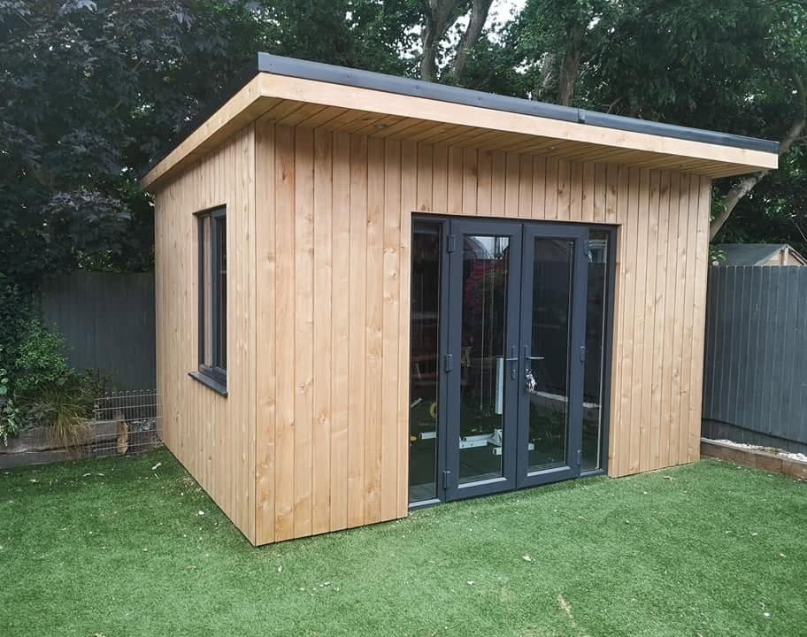 Finished timber-clad garden room with grey French doors and a flat roof overhang, standing on artificial grass