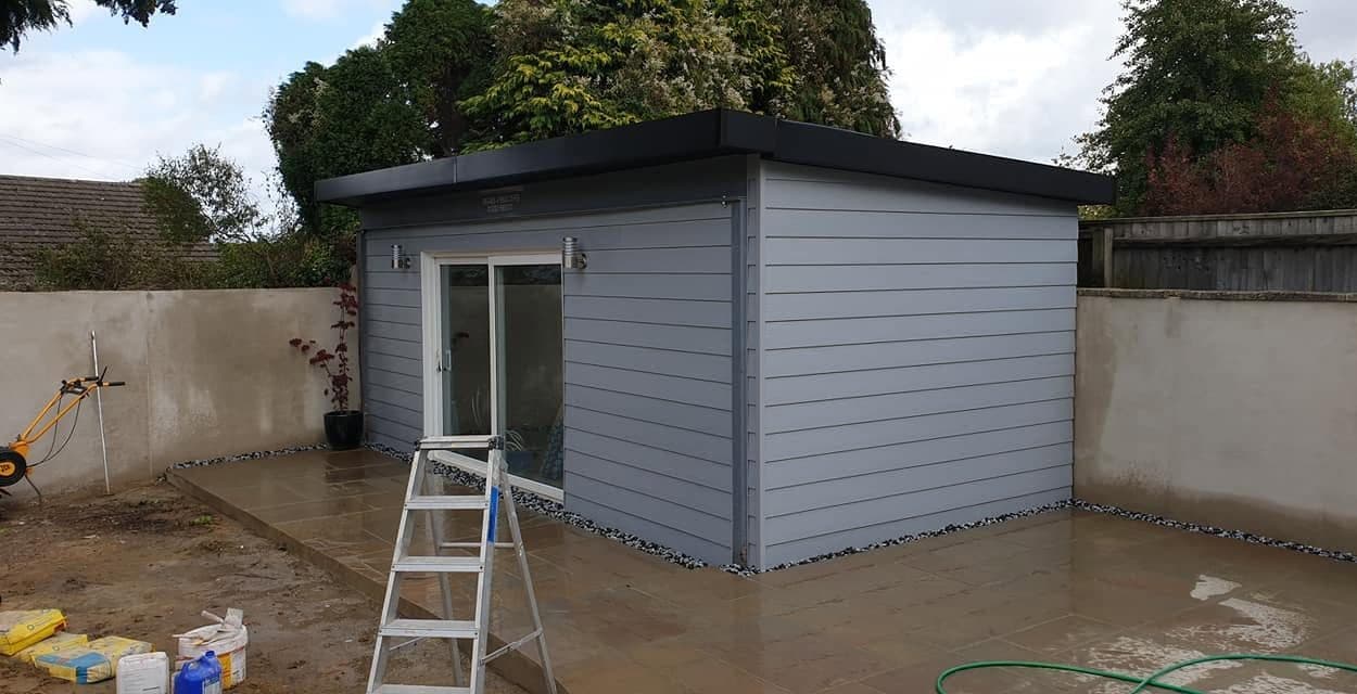 Grey-clad garden room with flat roof and sliding doors, new sandstone patio and chipping strip round its base