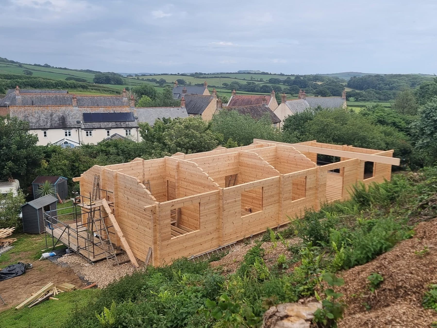 Log building walls up to gable height on a hillside plot, scaffold tower at one end, village houses behind