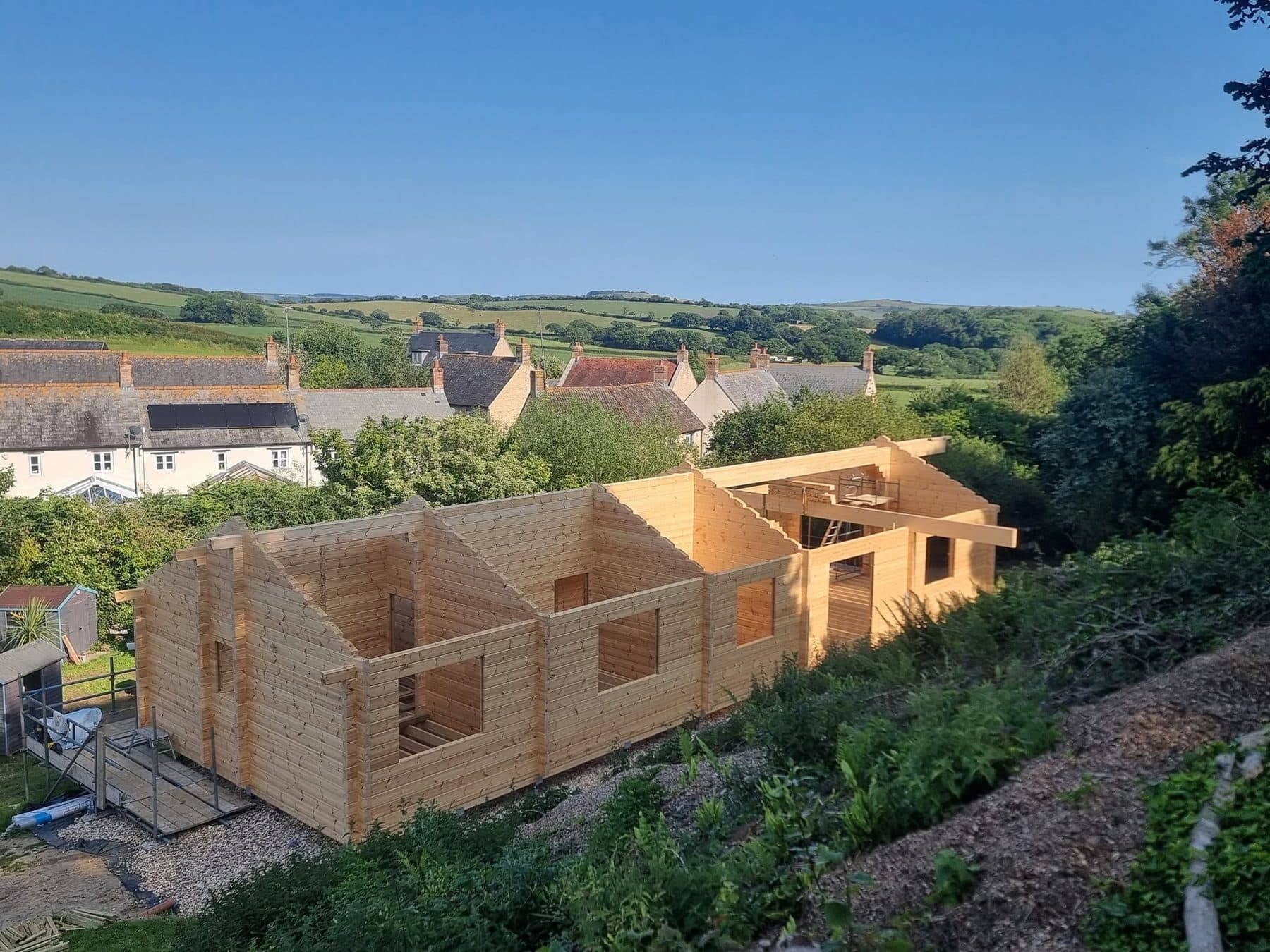 Log building shell in sunshine with gables up and ridge beam fitted, countryside and village behind