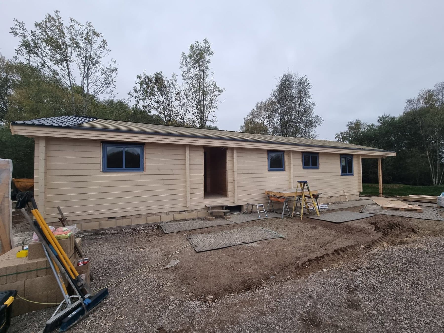Long log cabin with grey-blue windows on a block plinth, roof covering part fitted, site still in progress