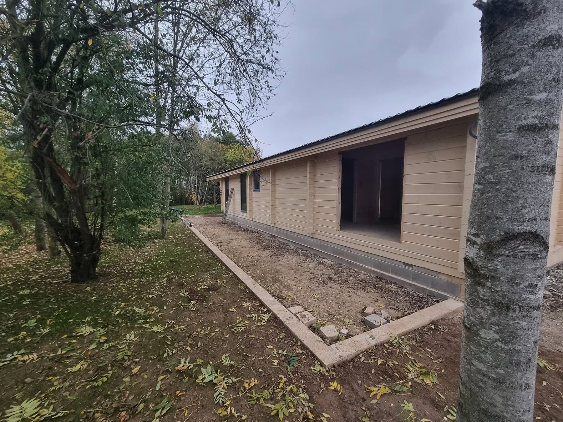 Side of a log cabin with a large door opening, dark profiled roof and a block-edged base strip alongside