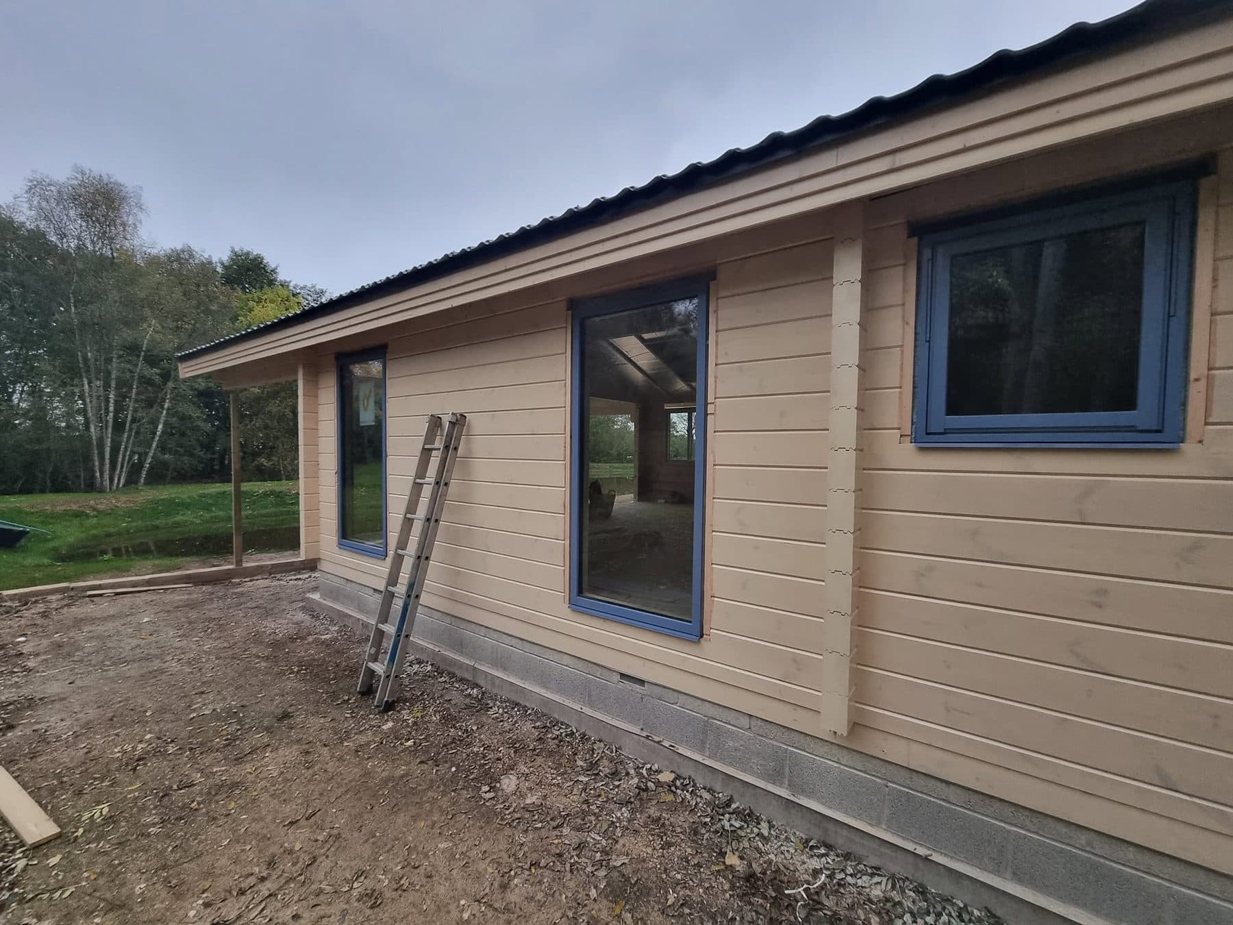 Log cabin wall with grey-blue framed windows on a blockwork plinth with an air brick, ladder against the wall