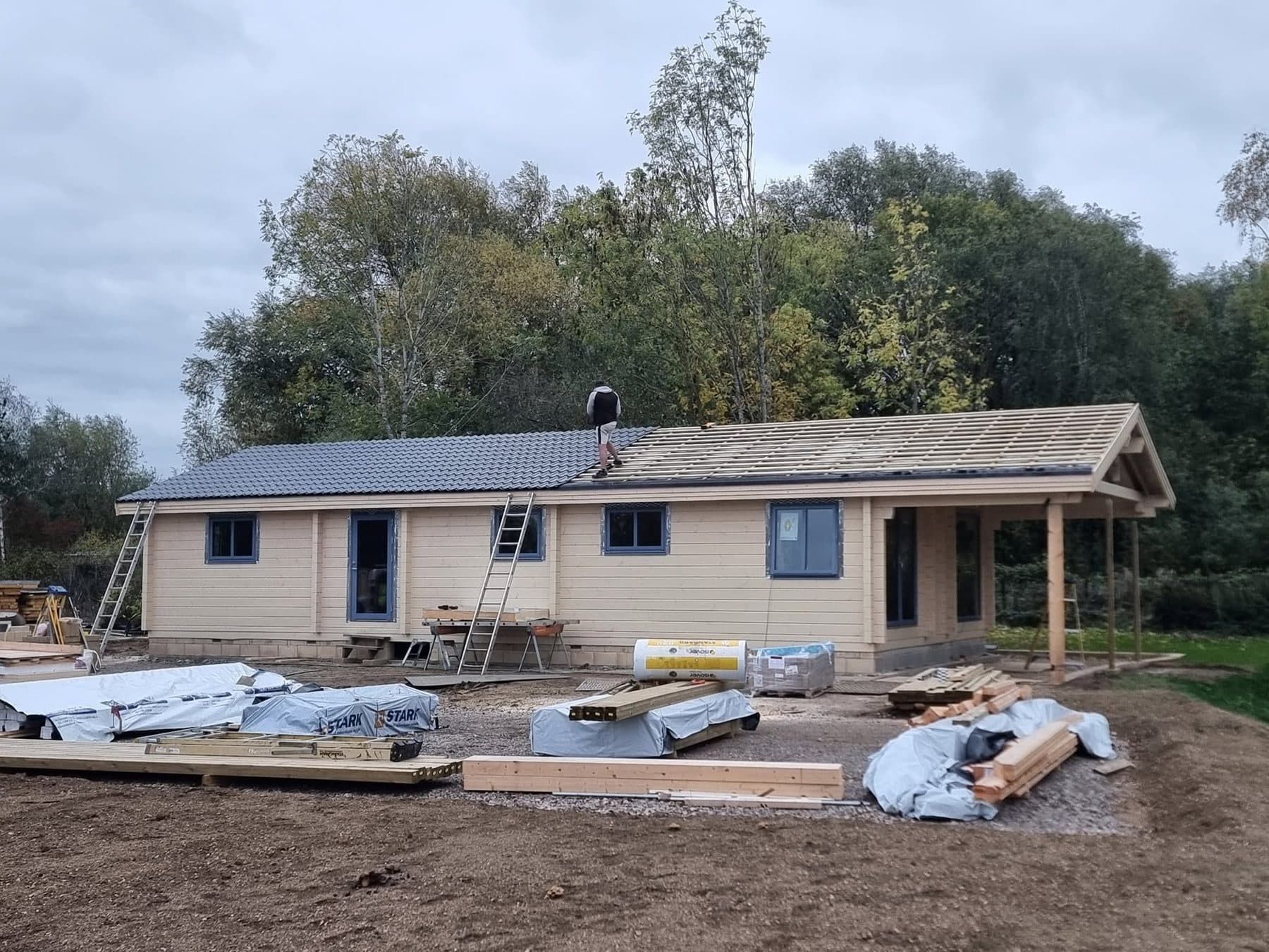 Log cabin with roof sheets going on over battens, covered porch at one end, materials stacked in front