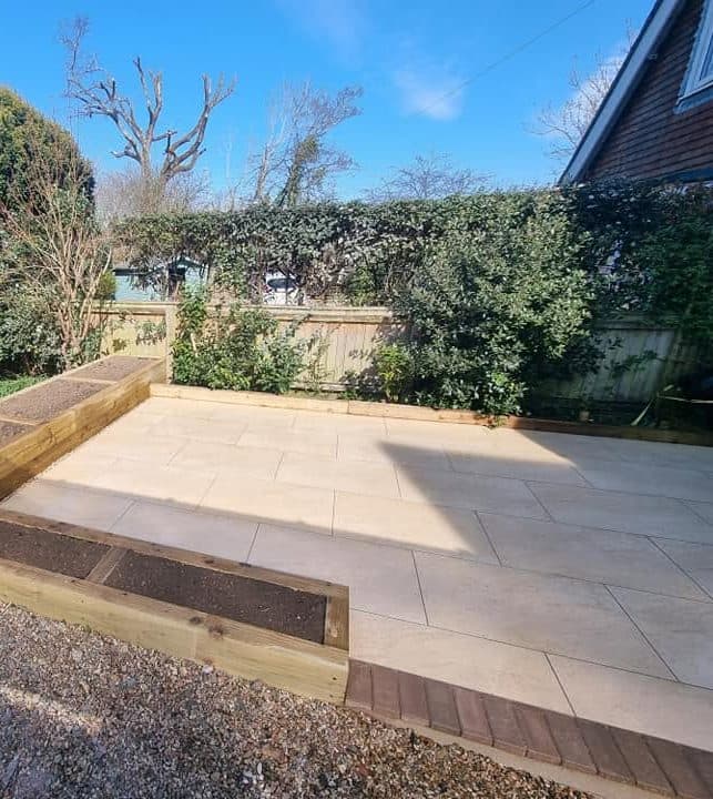 Pale paved patio with L-shaped timber sleeper raised beds and brick edging, set in gravel