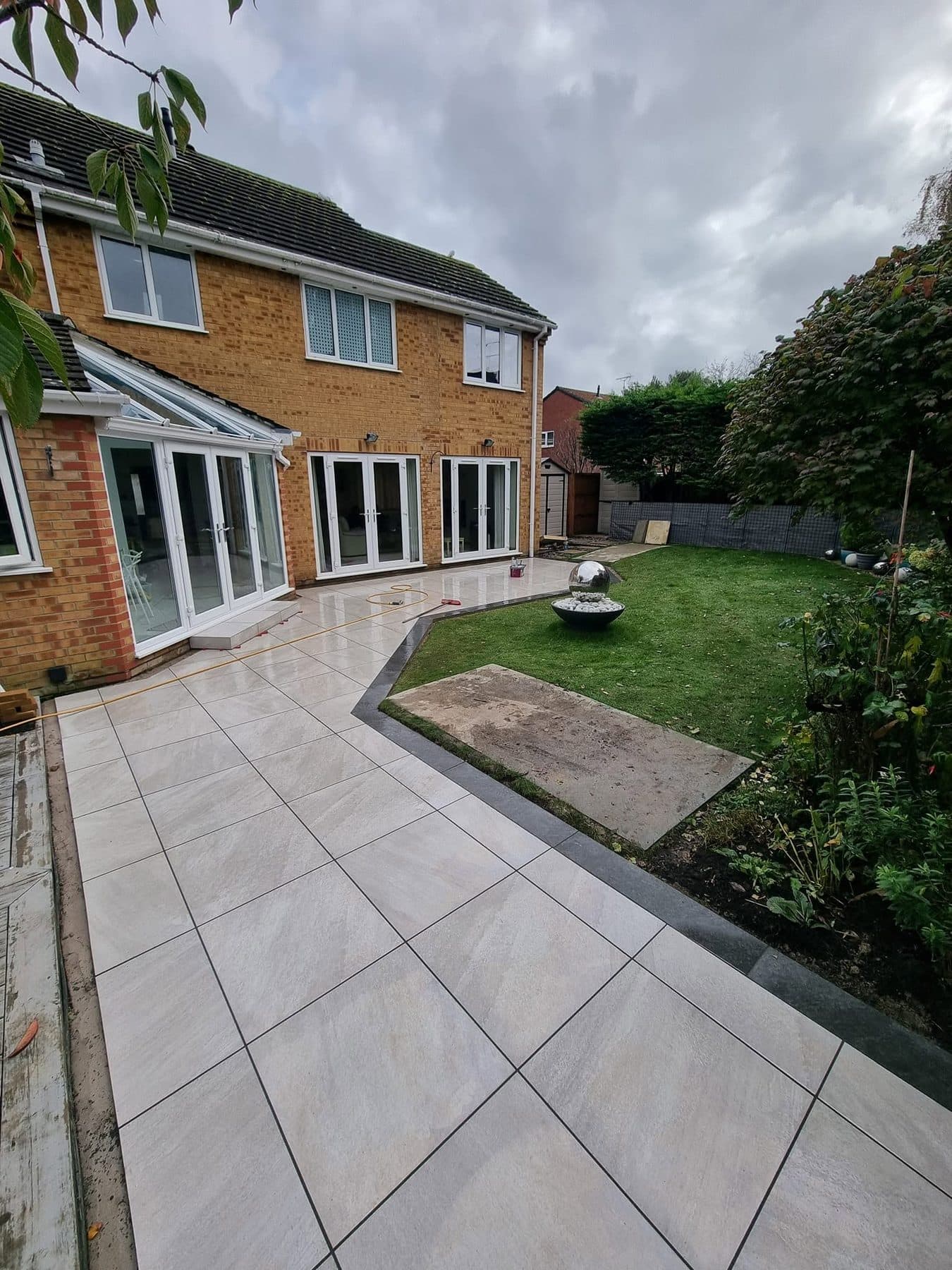 Large light grey porcelain patio with a dark border wrapping a lawn behind a brick house