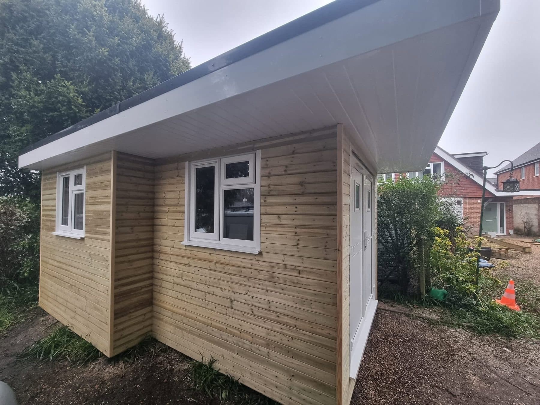 Timber-clad garden building with a wide overhanging flat roof, white uPVC windows and double doors