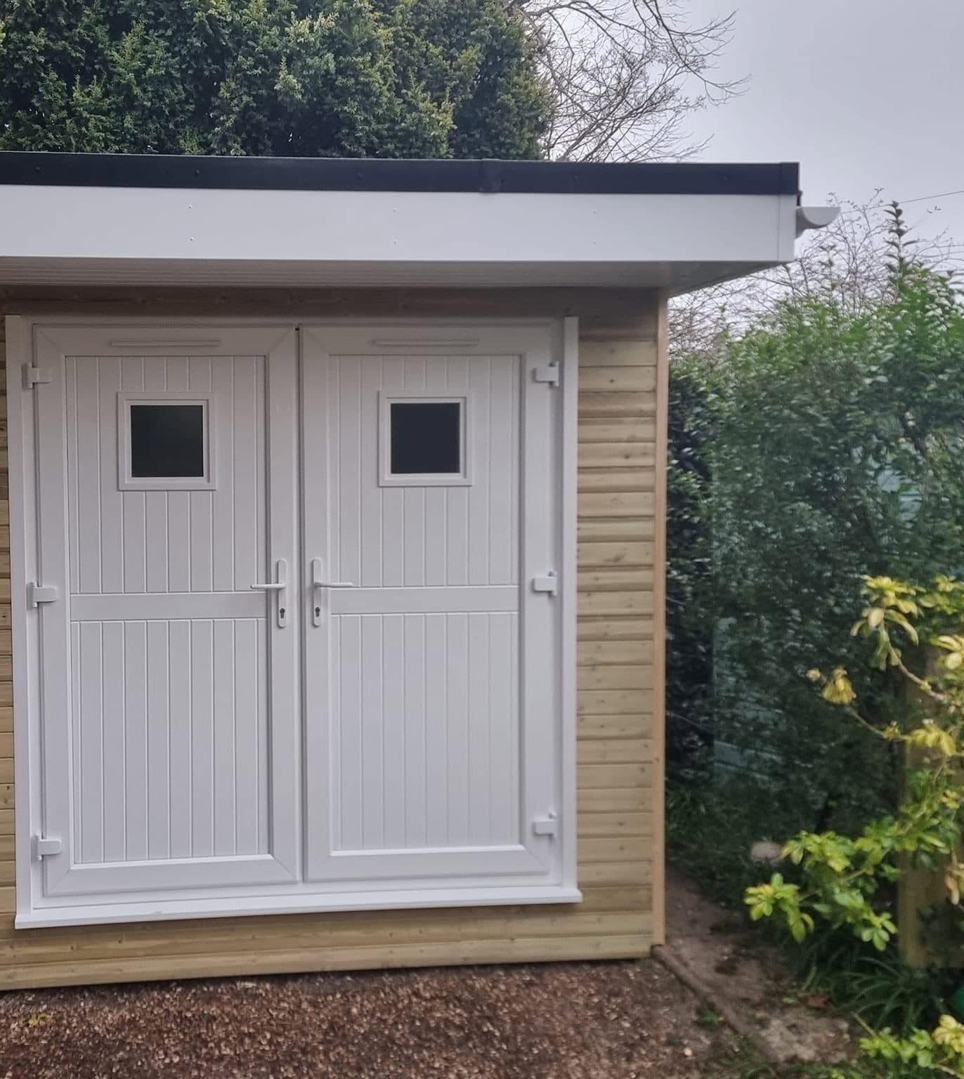 White uPVC double doors on a new timber-clad garden building with a flat roof and white fascia