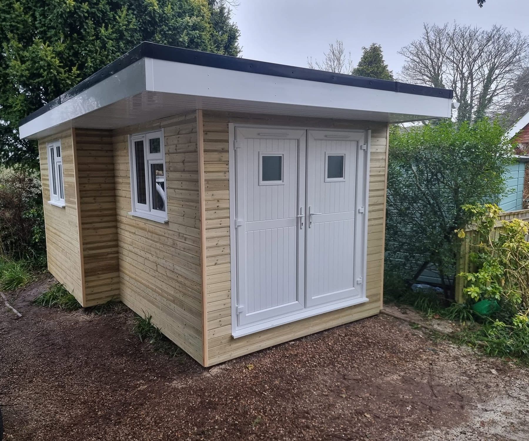 New timber-clad garden building with flat roof, white fascia, uPVC windows and double doors on a gravel drive